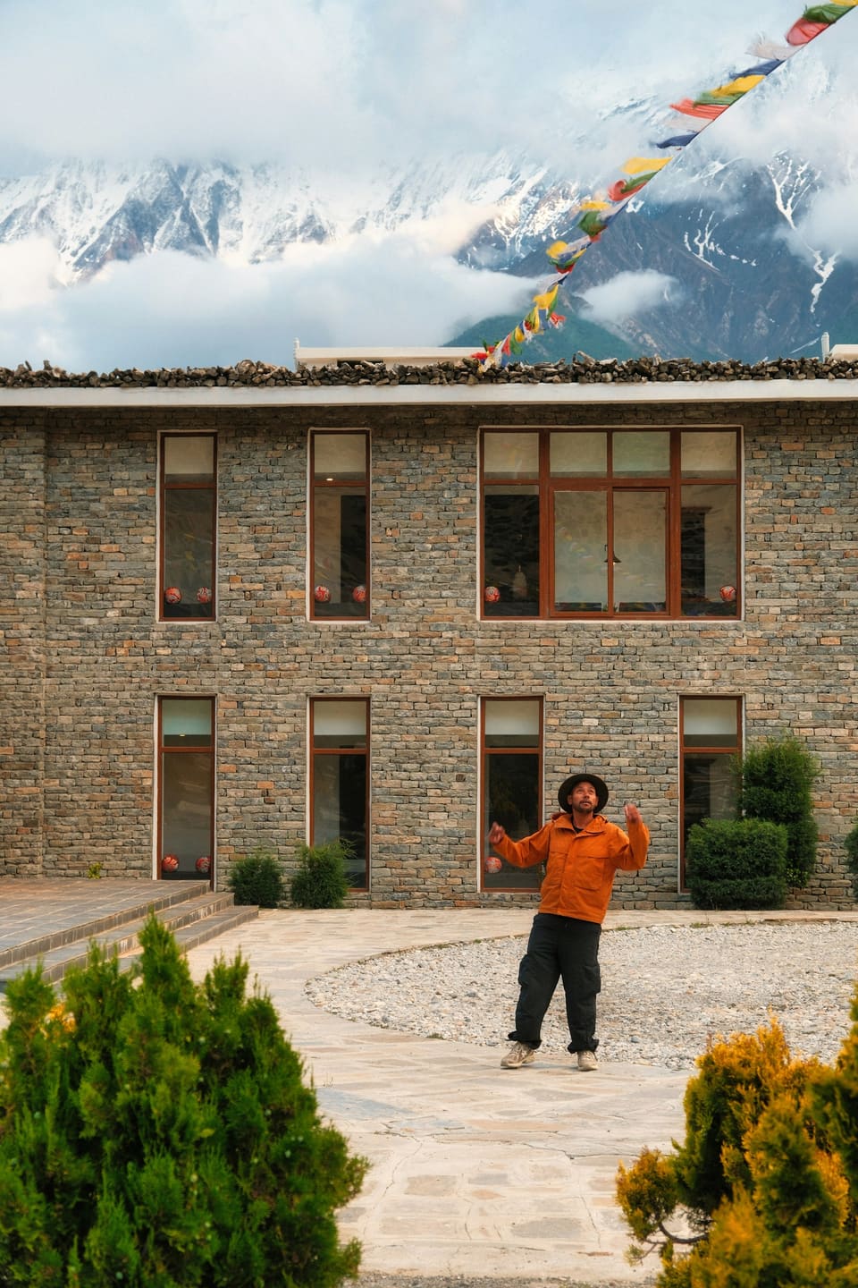A young man in an orange jacket outside a stone building in the hills